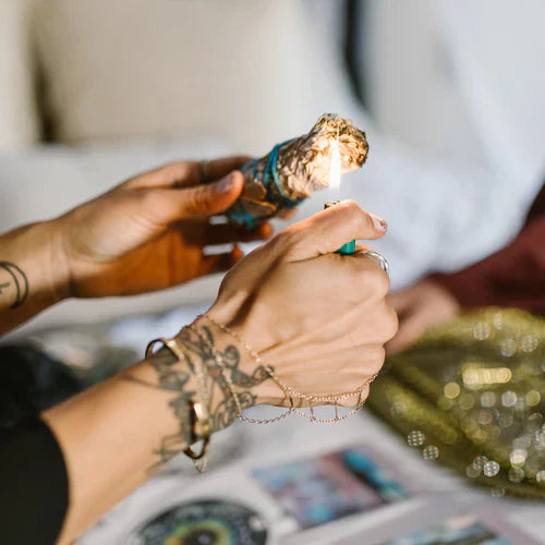 Hands lighting a tied herb bundle beside candles on a table.