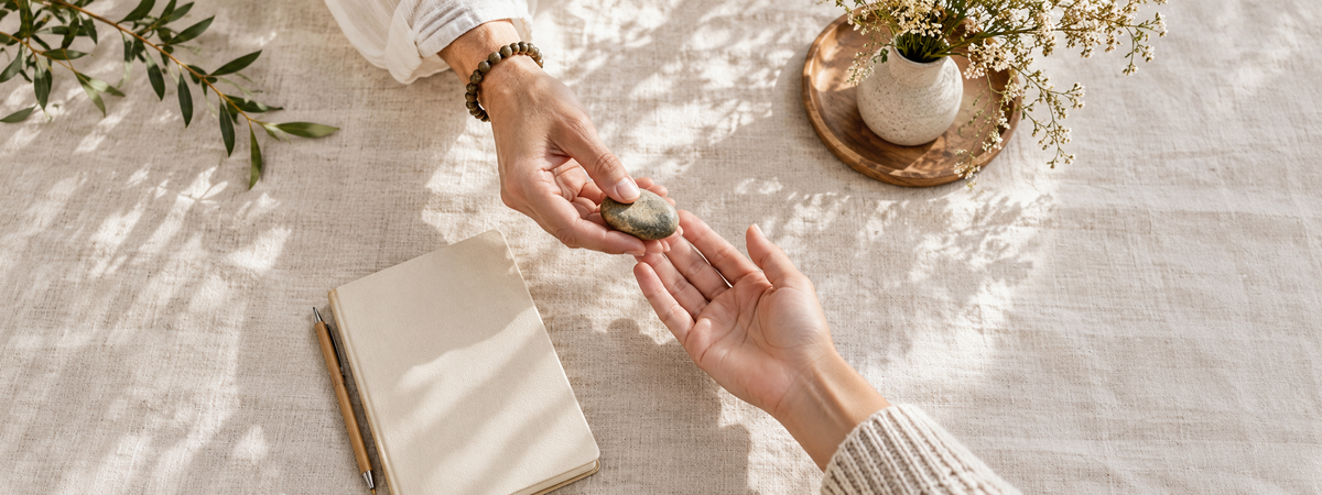 Two adult hands exchanging a small plain stone across a pale linen surface, with a blank notebook nearby.