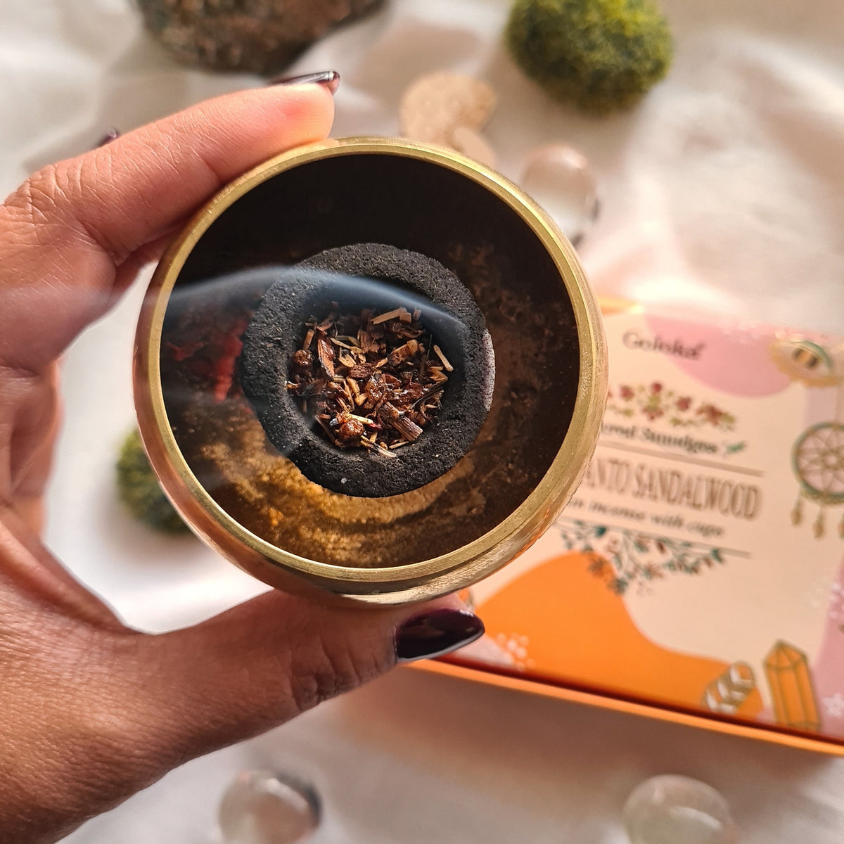 Hand holding a brass-coloured incense bowl containing charcoal and resin.