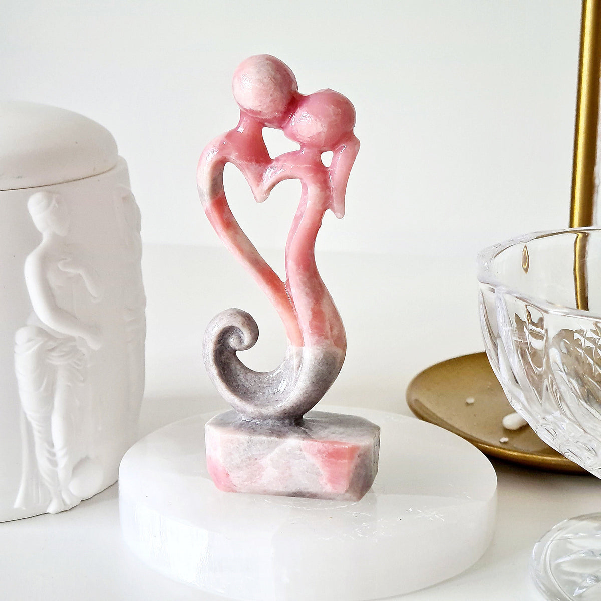Pink and grey carved figure sculpture displayed on a white stand beside a glass vessel.