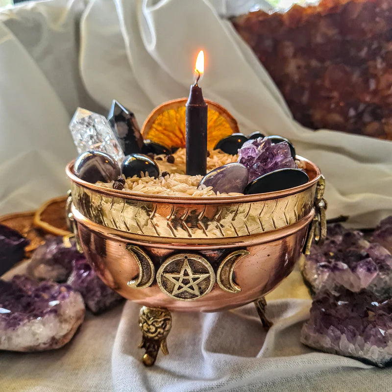 Brass-coloured ritual bowl with a triple moon motif beside crystals and a lit candle.