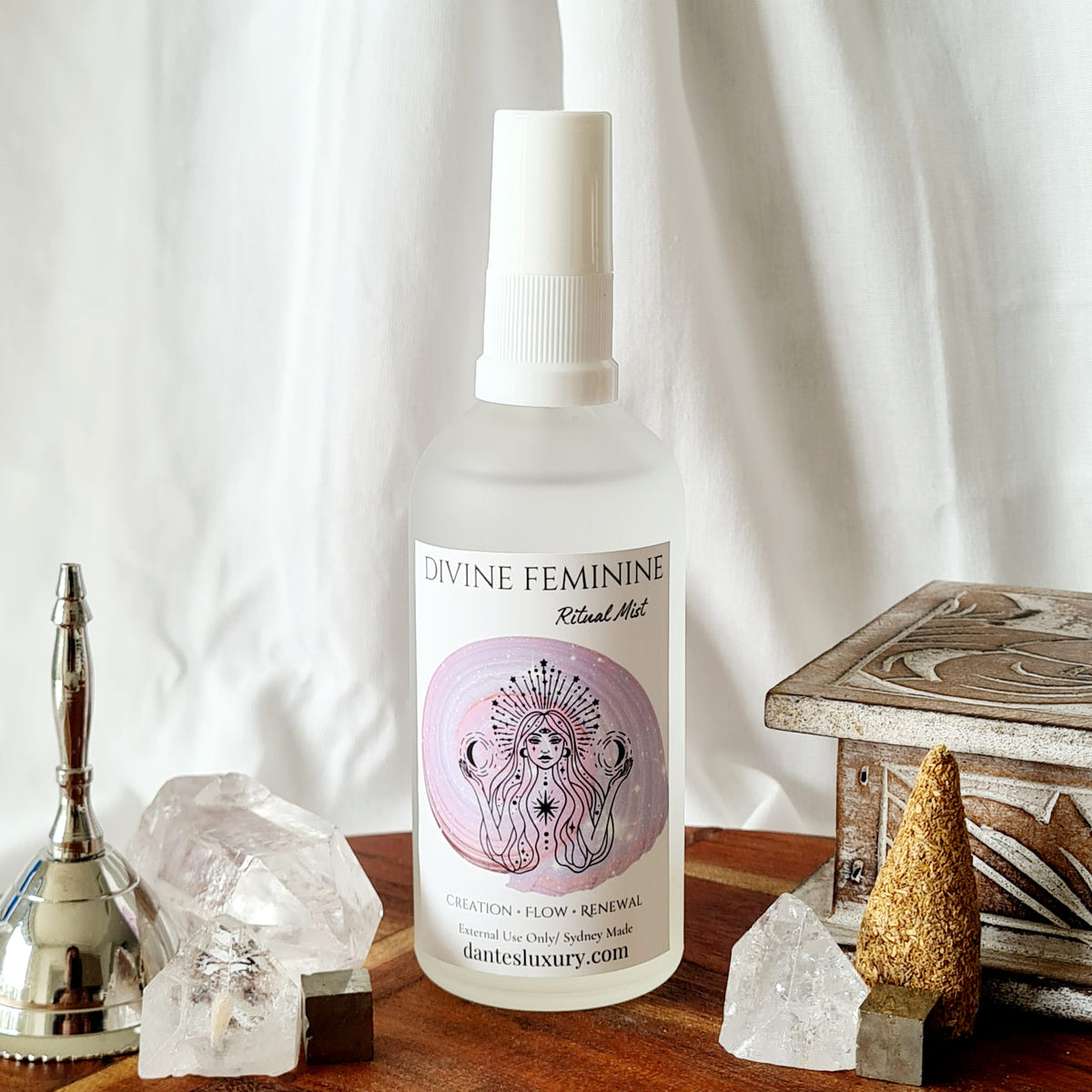 Divine Feminine ritual mist bottle with a pink illustrated label, arranged beside quartz crystals and a brass bell.