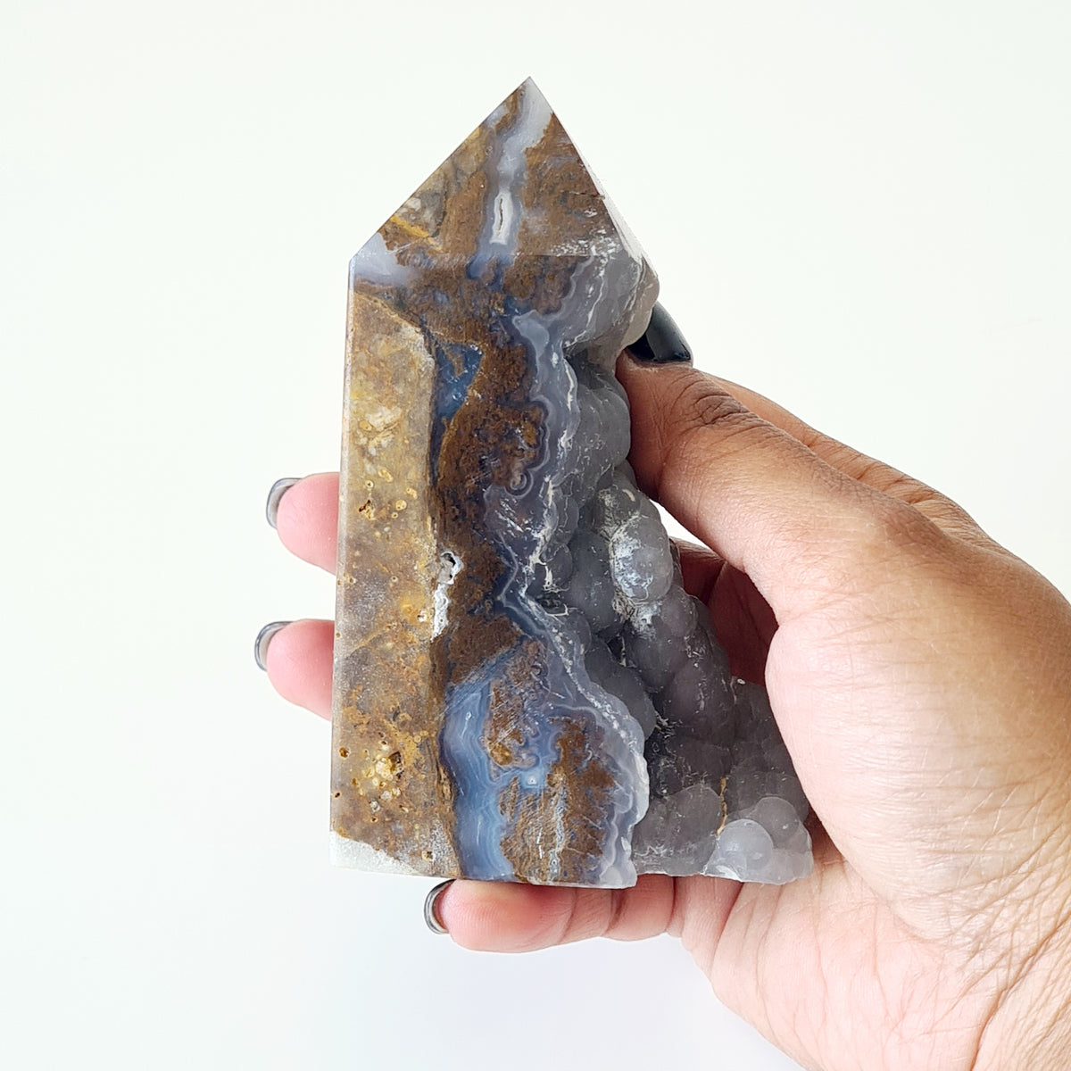 Hand holding a botryoidal agate tower with a natural pocket, blue-grey bands and golden-brown areas.