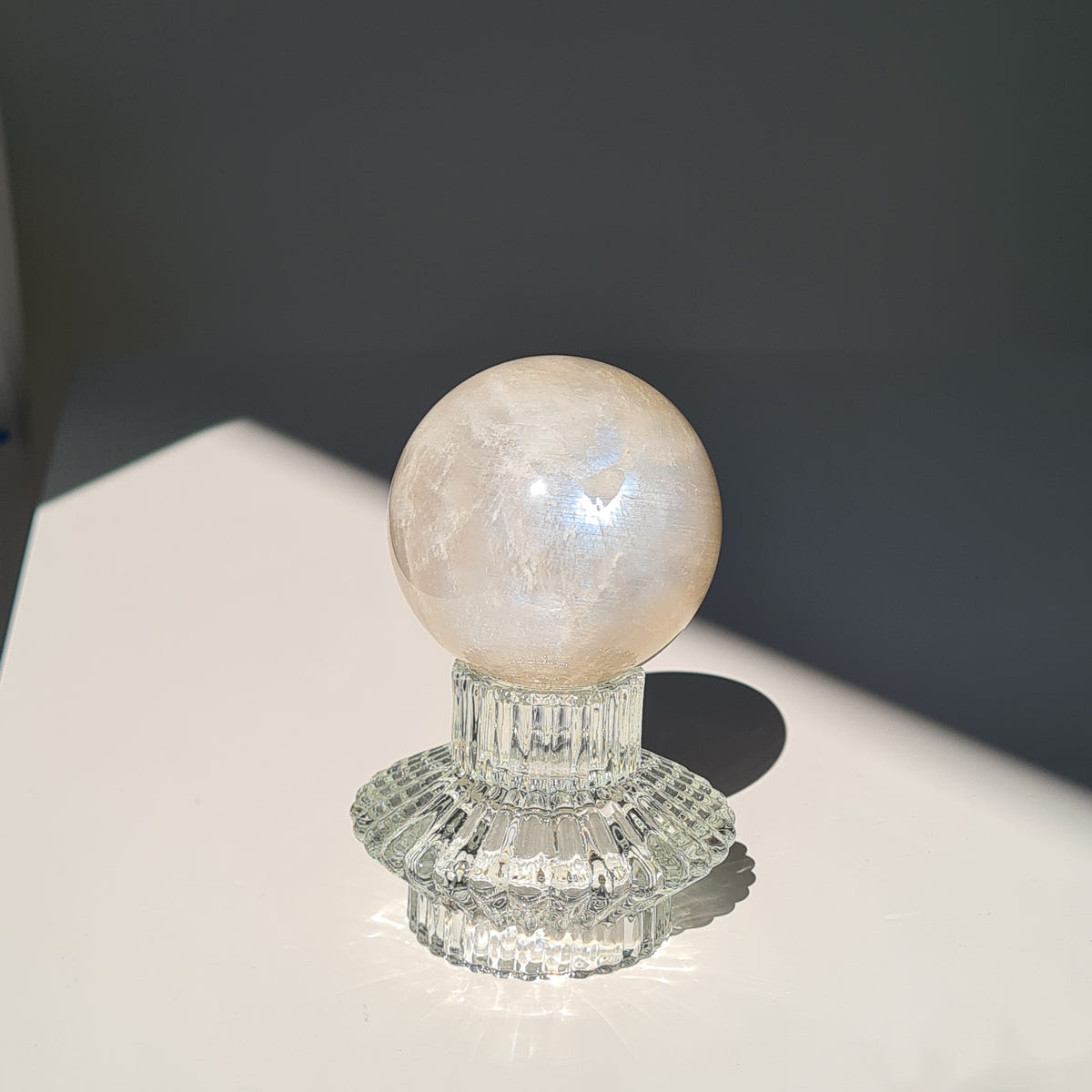 Moonstone sphere H with a warm cream surface and a pale blue flash on a short clear decorative stand.