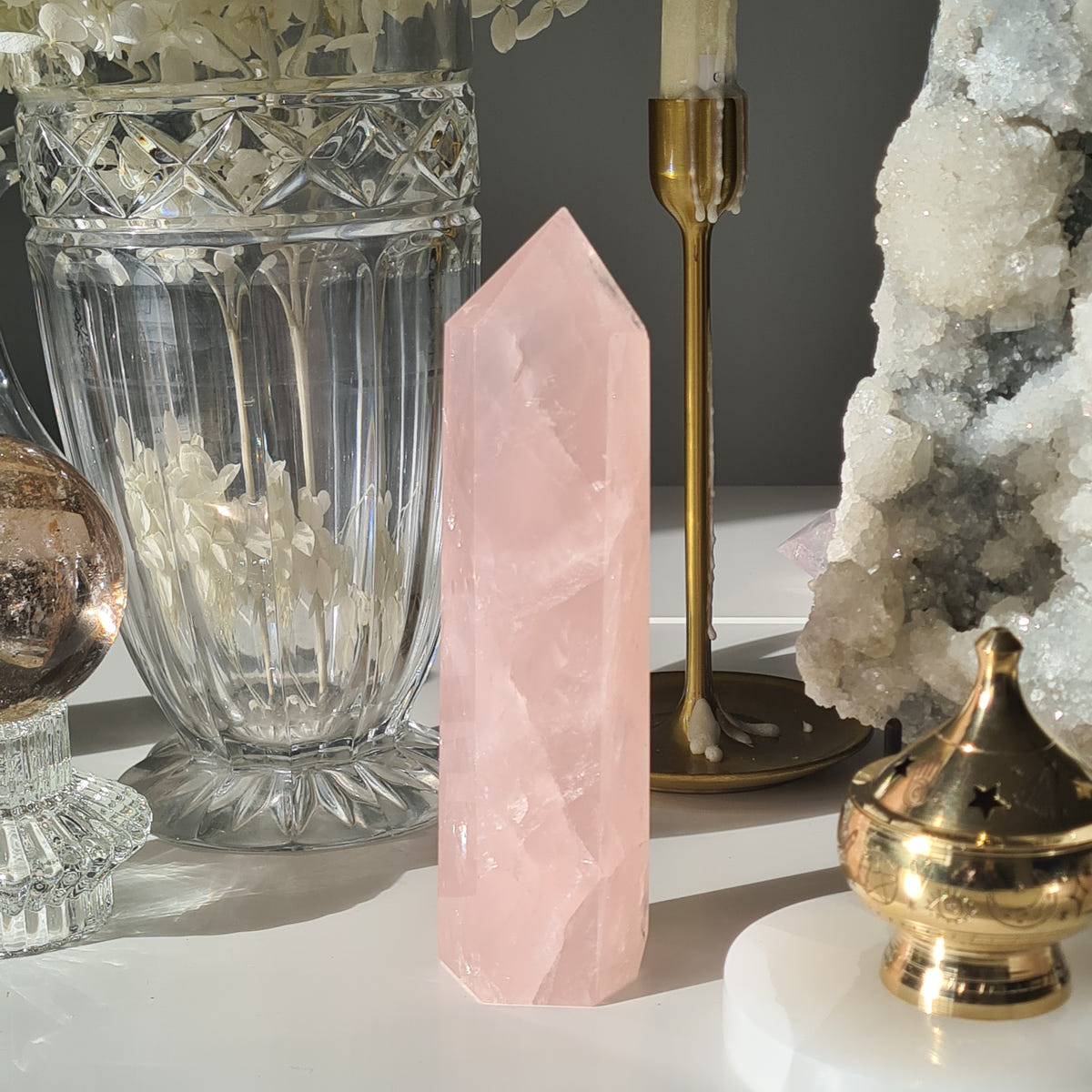Tall pale pink rose quartz point with cloudy diagonal bands, displayed beside glassware and a brass burner.