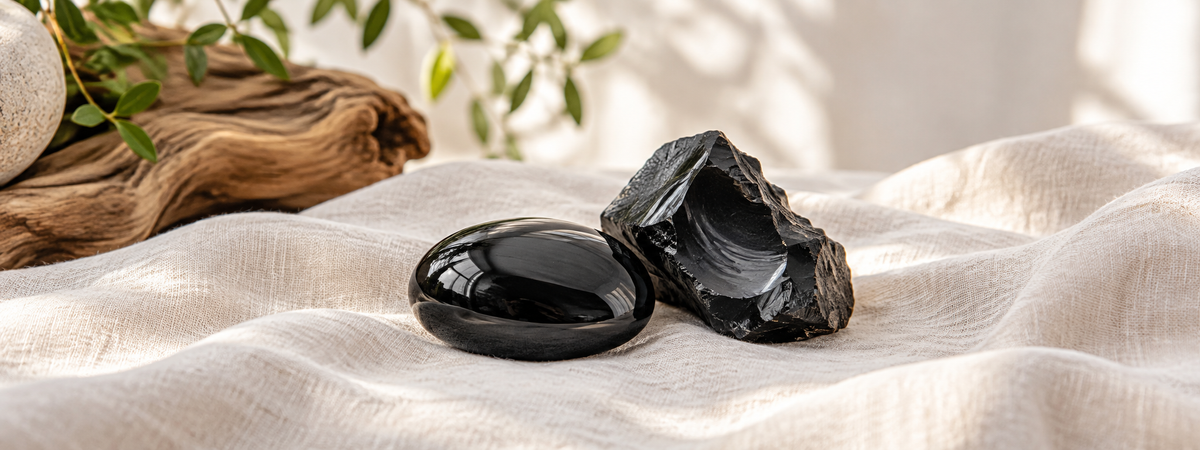 A smooth polished black obsidian palm stone beside an unpolished dark volcanic-glass piece on ivory linen.