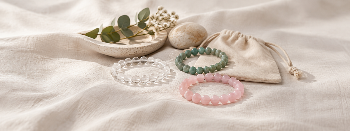 Three modest bead bracelets in clear, soft pink and muted green tones resting on ivory linen beside a plain storage pouch, whole bracelet outlines visible.