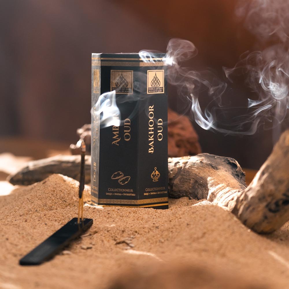 Black and gold oud incense box beside a lit incense stick with rising smoke in a sandy display.