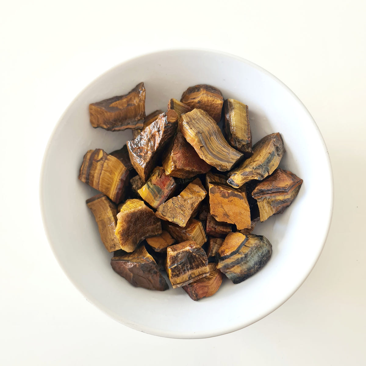 Small rough tiger’s eye pieces with golden-brown and dark bands arranged in a white bowl.
