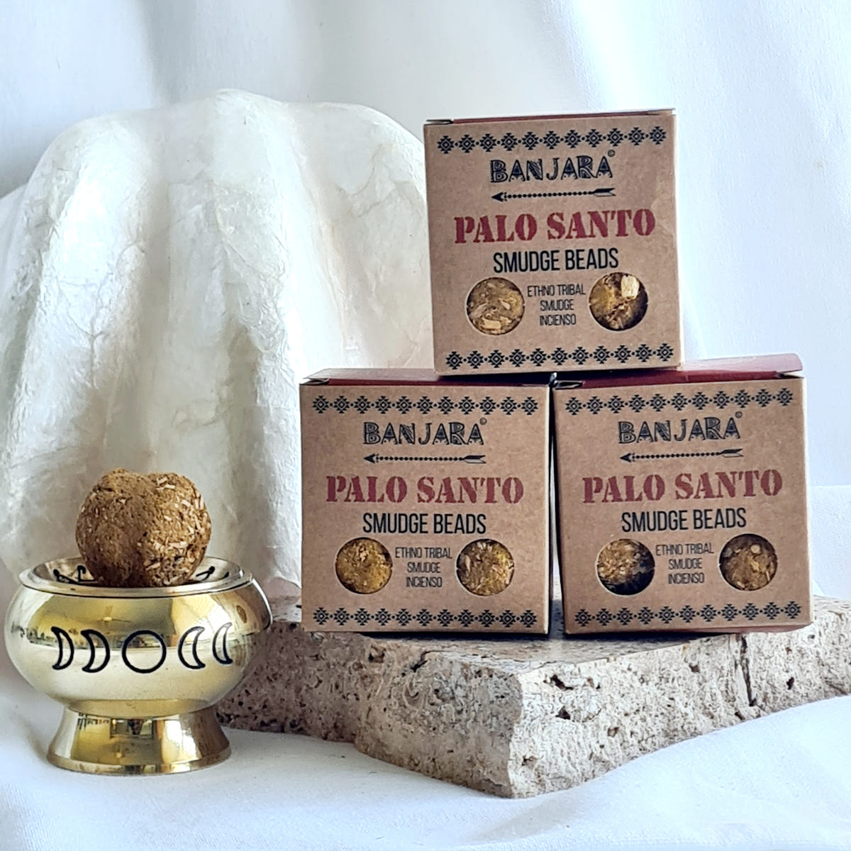 Three Banjara Palo Santo smudge bead boxes beside a brass moon-design burner holding a brown round bead.
