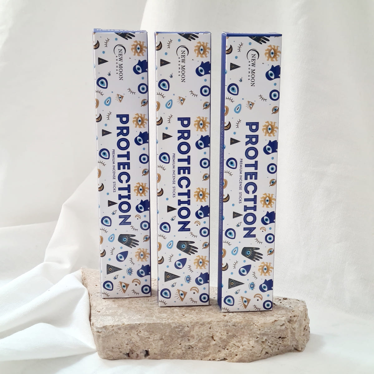 Three white Protection incense boxes with blue evil eye, moon and star motifs on a stone display block.