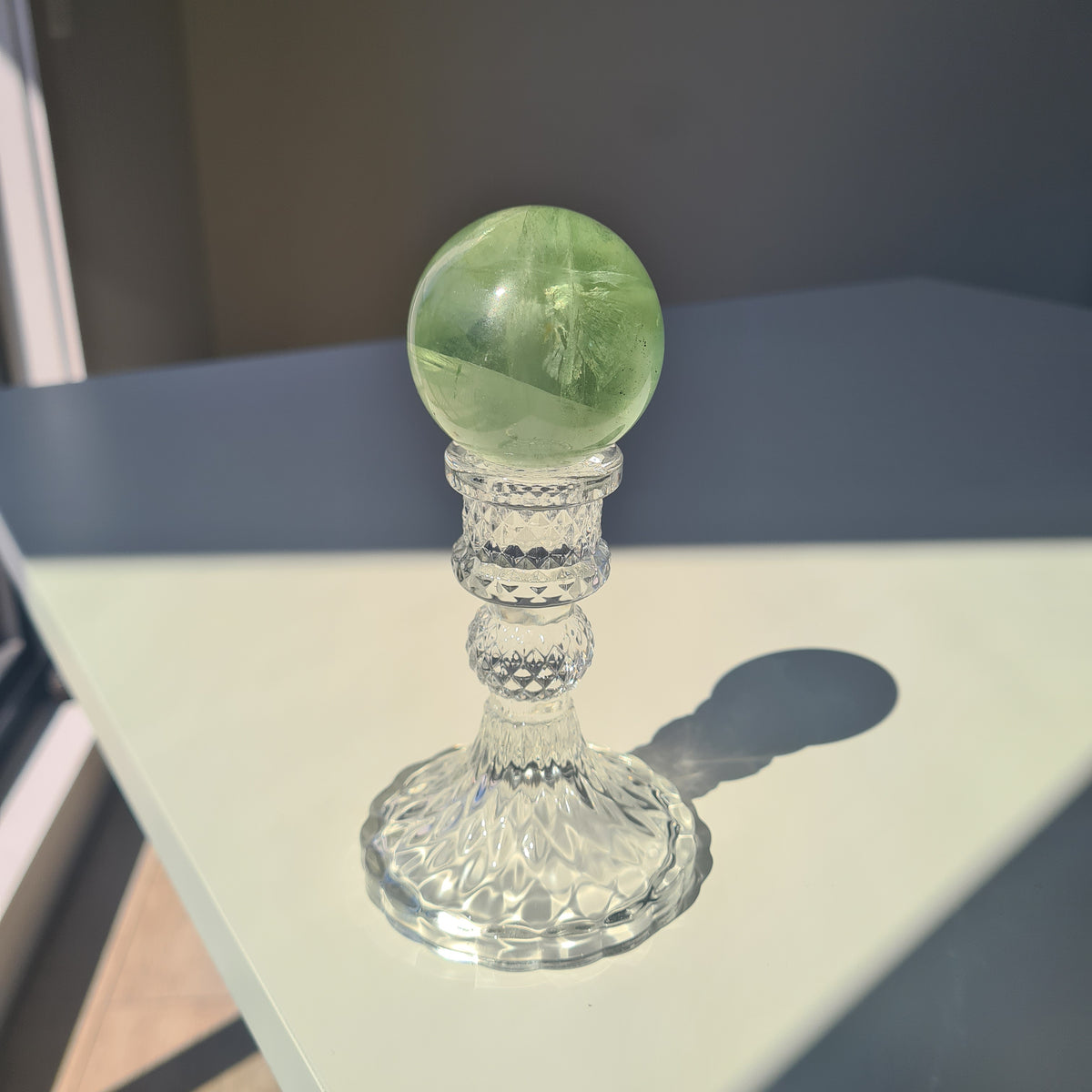 Green fluorite sphere with pale natural bands on a clear decorative stand in sunlight.