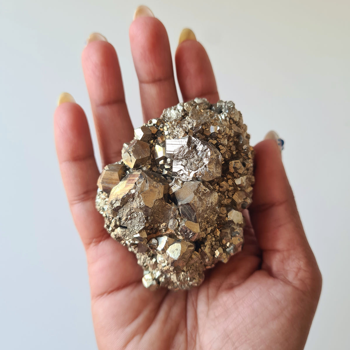 Hand holding pyrite specimen L with broad angular crystals across a rounded mineral cluster.
