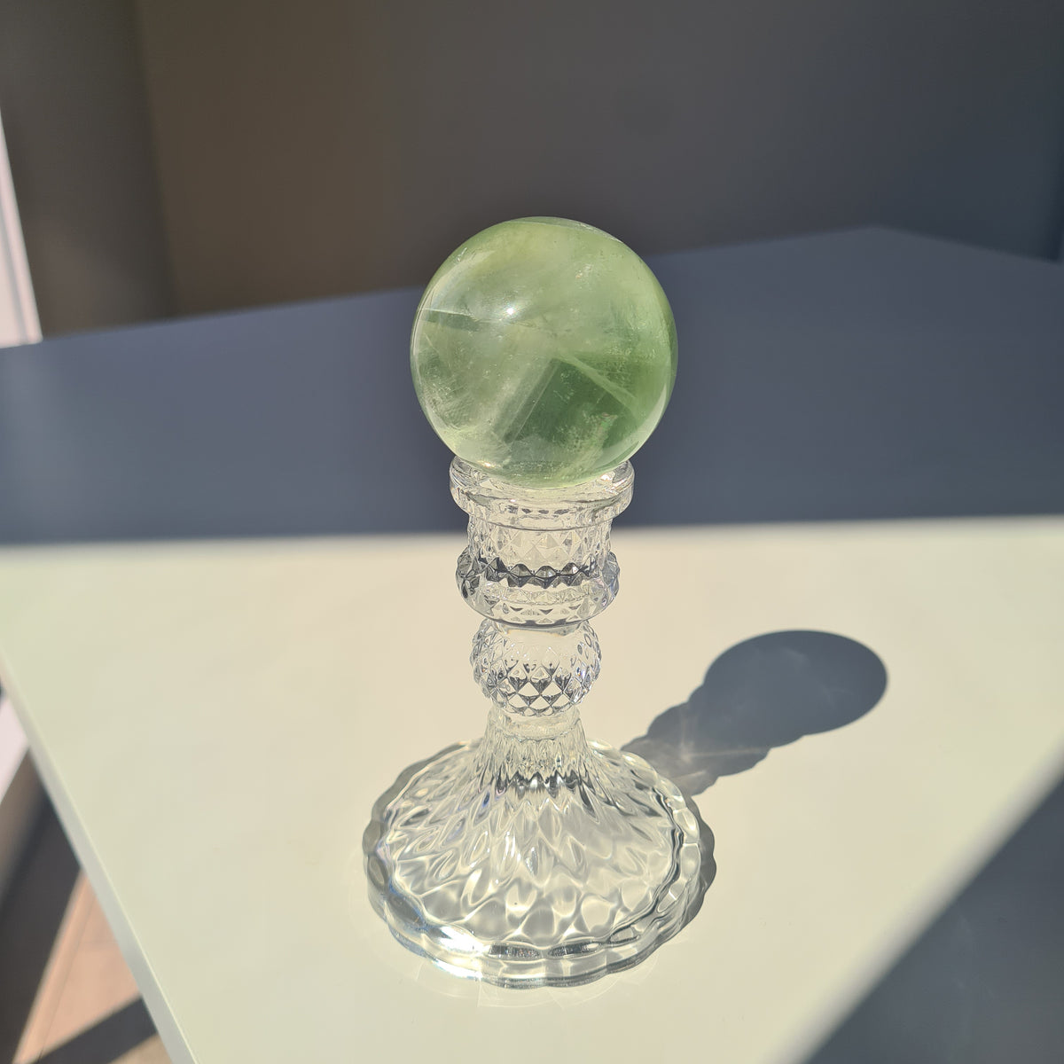 Green fluorite sphere with pale natural bands on a clear decorative stand in sunlight.