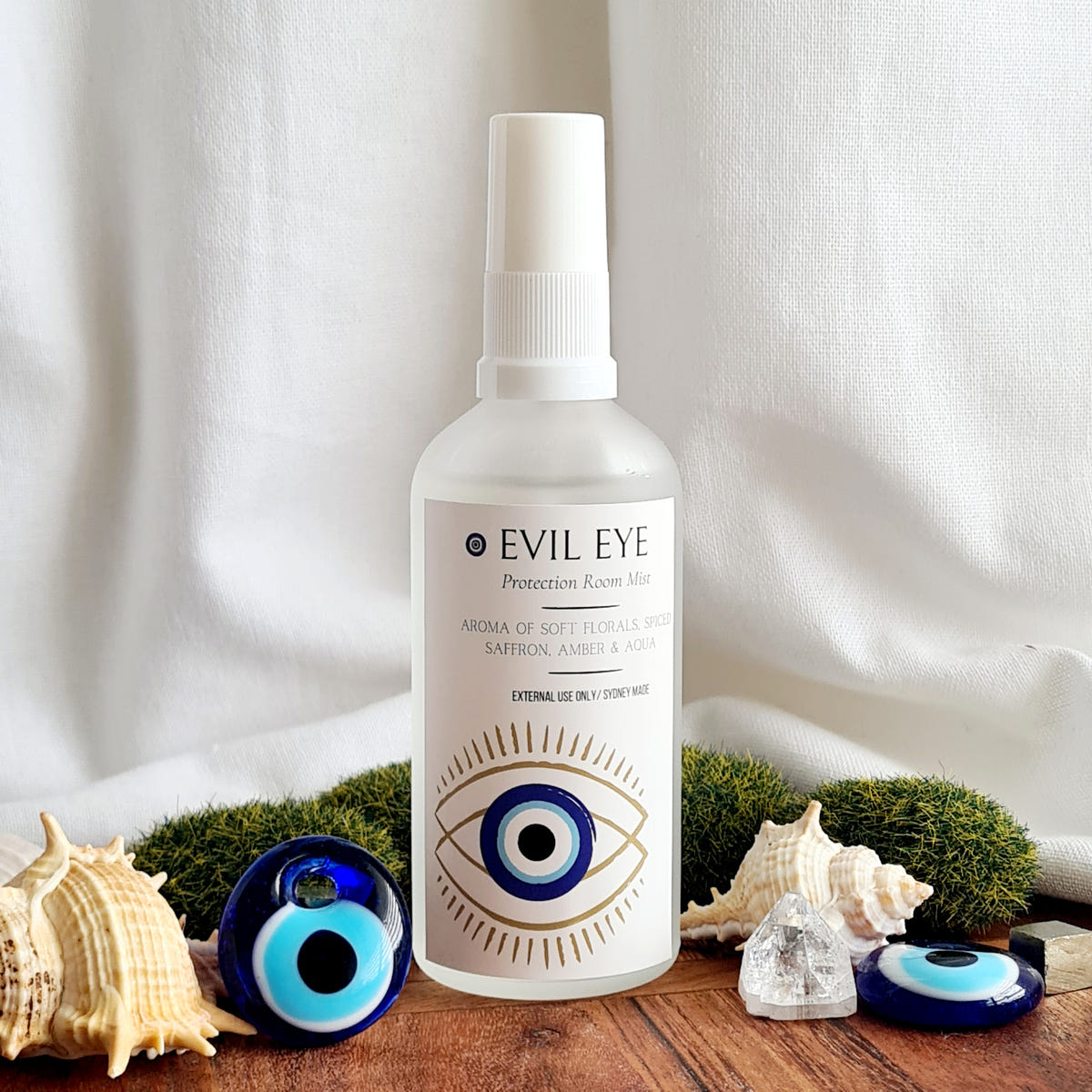 White Evil Eye ritual mist bottle with a blue eye design, arranged beside shells and blue eye charms.