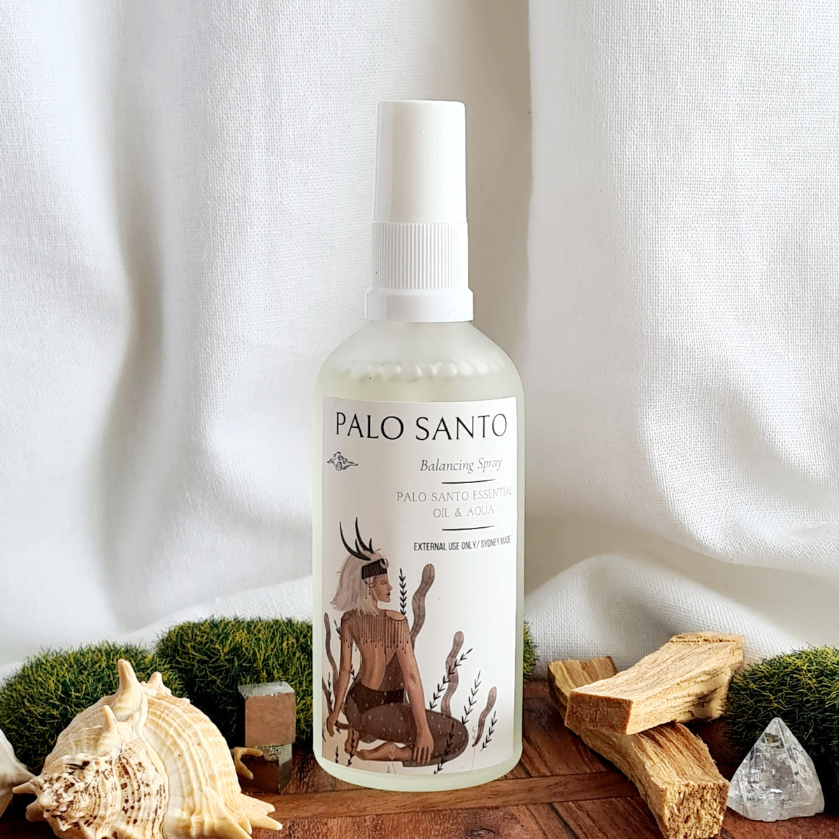 Palo Santo ritual mist bottle with an illustrated label, arranged beside wood sticks, a shell and a quartz crystal.