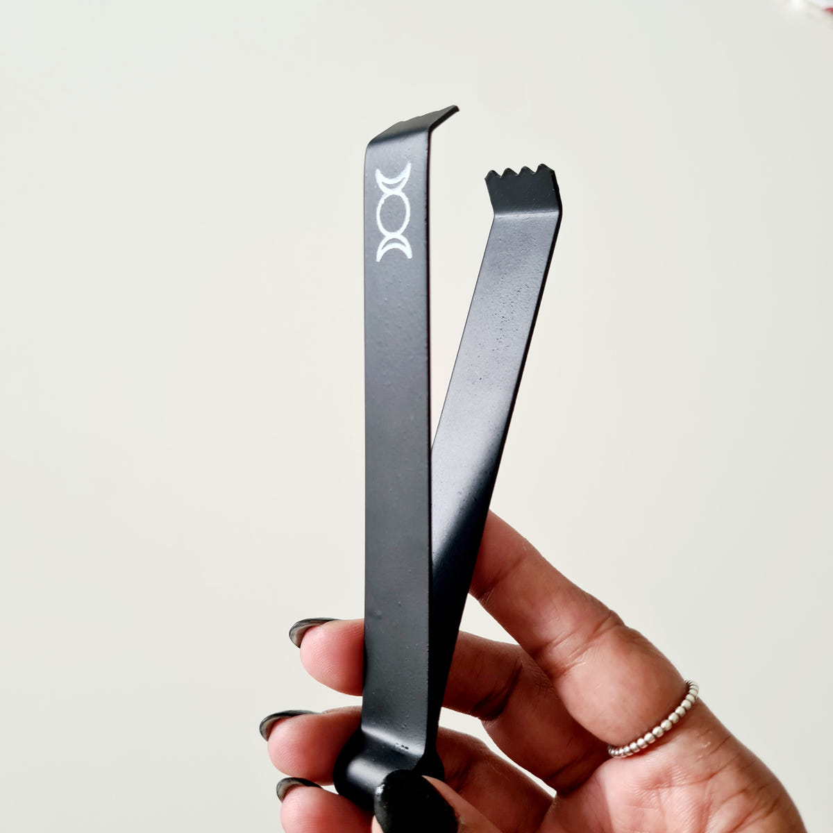Hand holding black incense tongs with serrated tips and a white triple moon symbol.