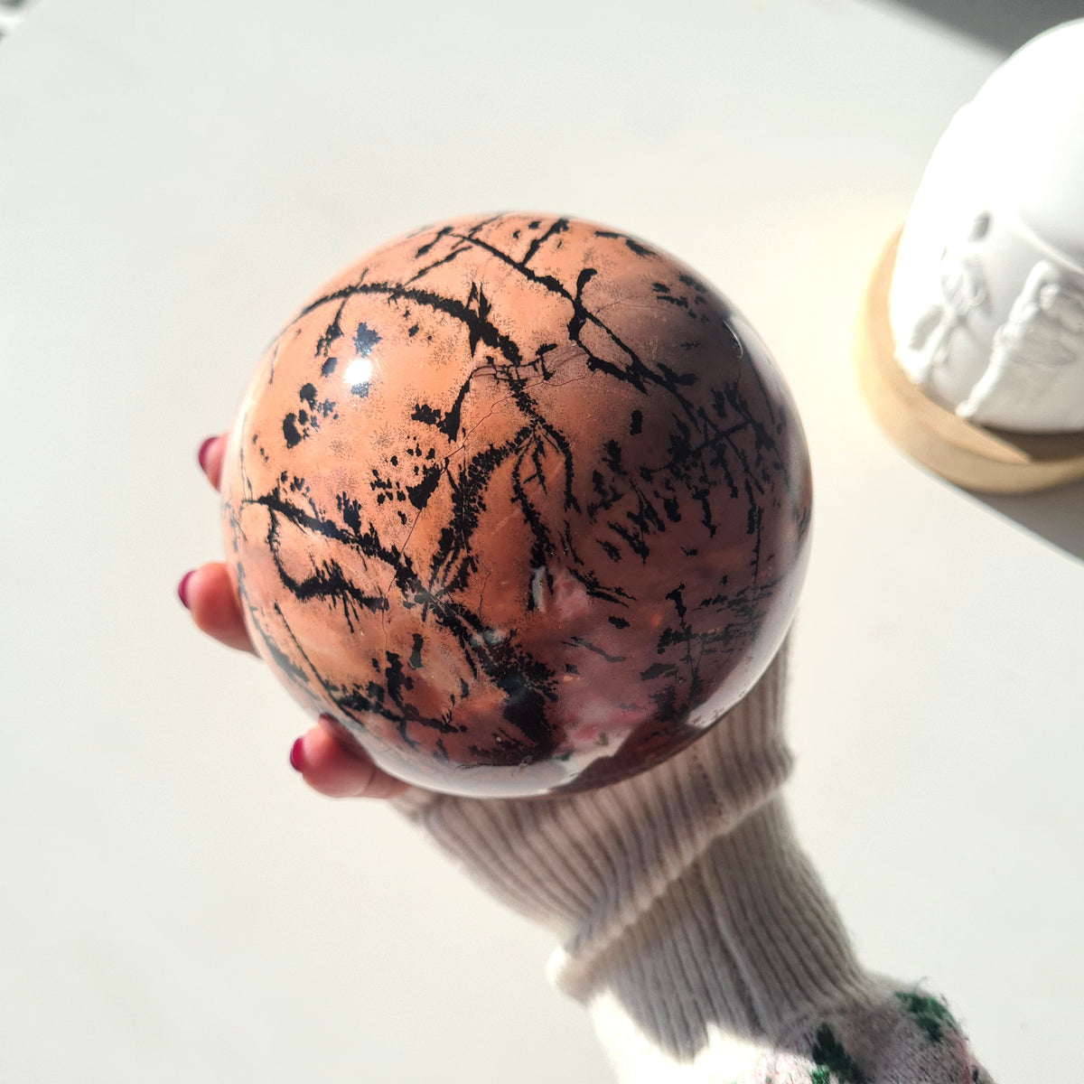 Close view of a picture jasper sphere in one hand, showing branching black patterns over peach tones.