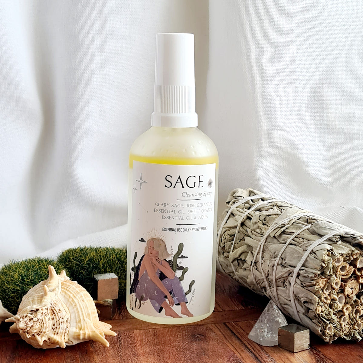 Sage ritual mist bottle with an illustrated label beside a tied sage bundle, a shell and quartz crystals.