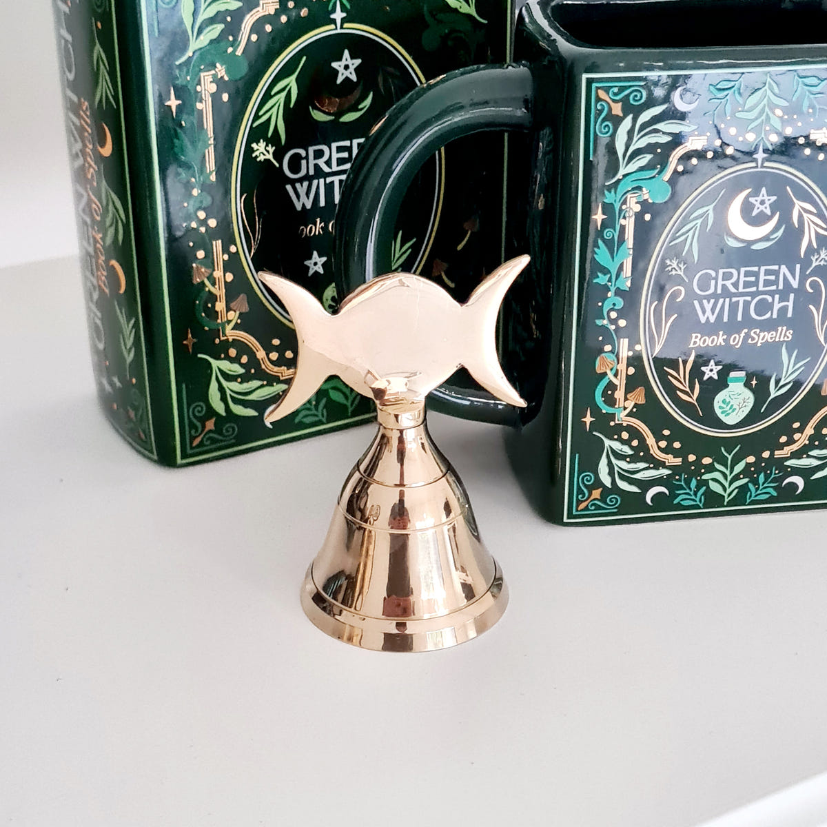 Gold-toned Triple Moon altar bell with crescent-shaped handle, displayed in front of books.