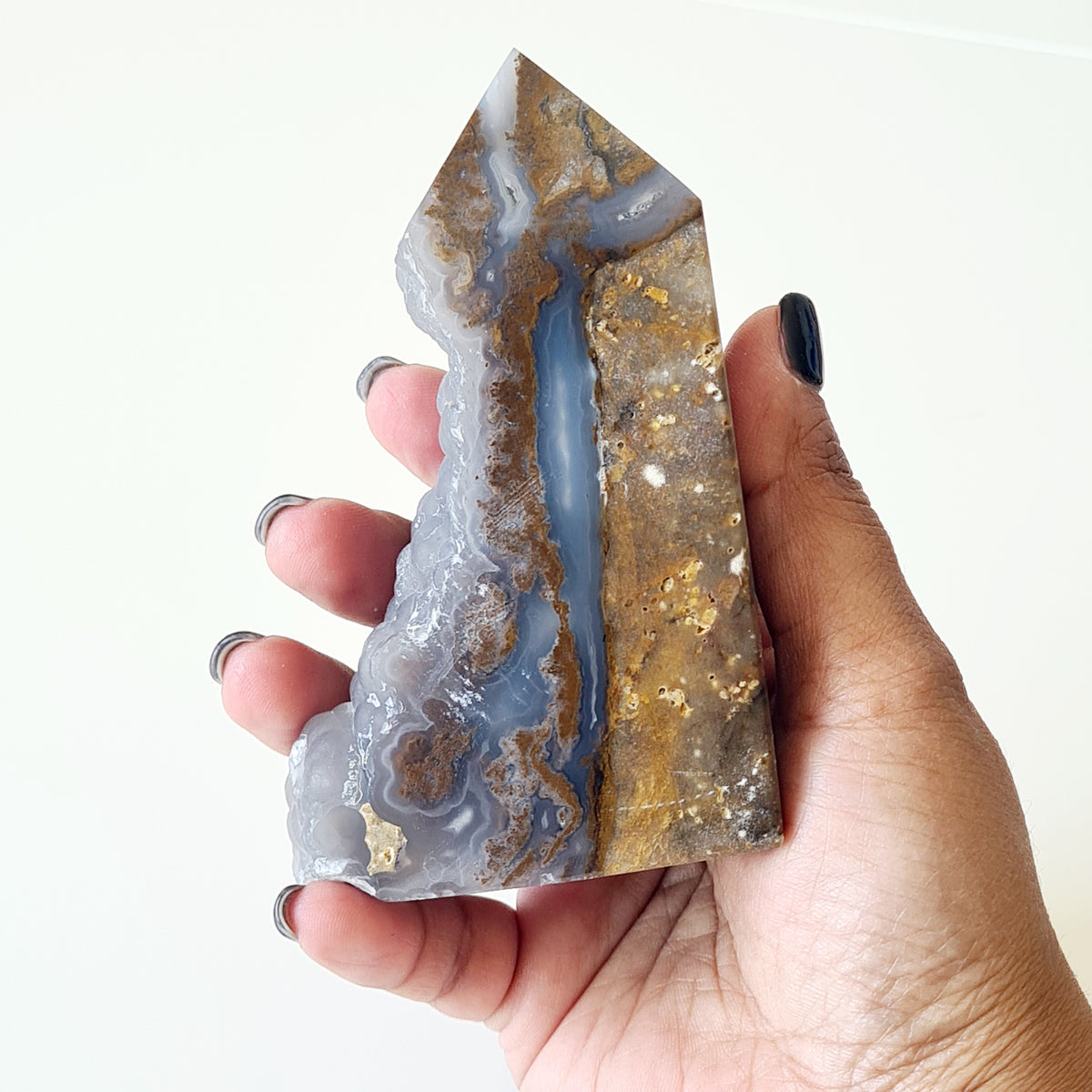 Hand holding a botryoidal agate tower with a natural pocket, blue-grey bands and golden-brown areas.