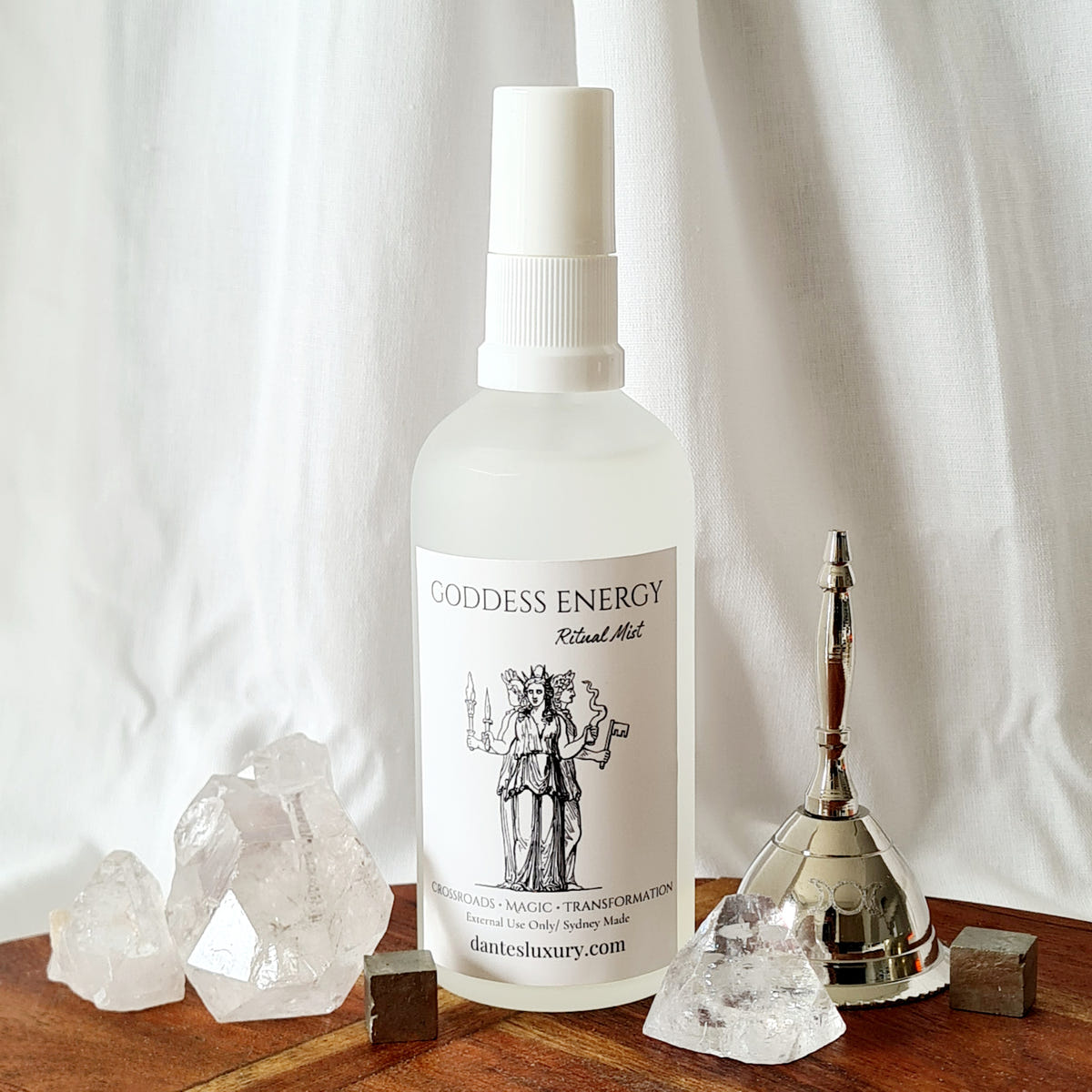 Goddess Energy ritual mist bottle with a black illustrated label, arranged beside quartz crystals and a brass bell.