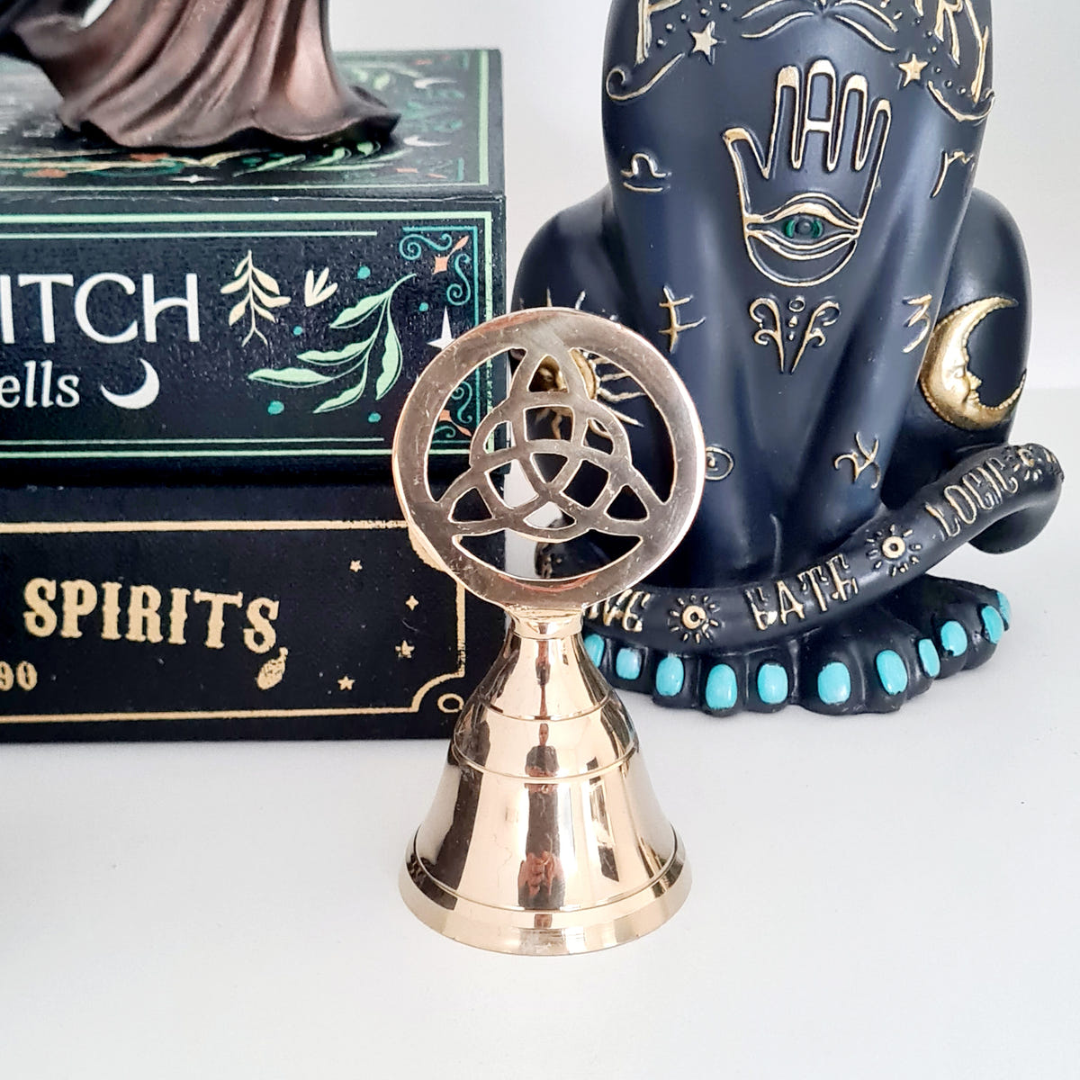 Gold-toned Triquetra altar bell displayed in front of books.