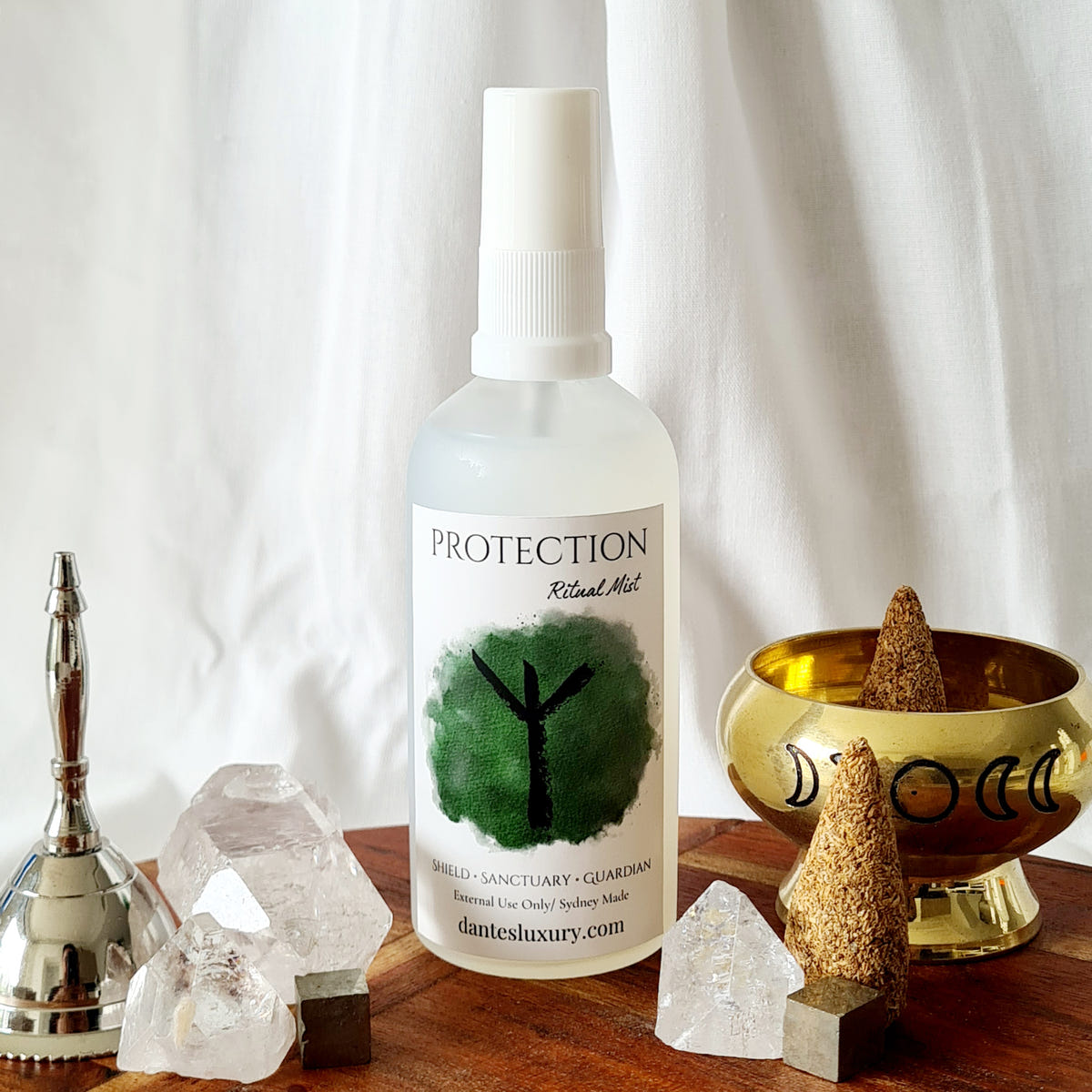 Protection ritual mist bottle with a green symbol label, arranged beside quartz crystals, a bell and a brass bowl.