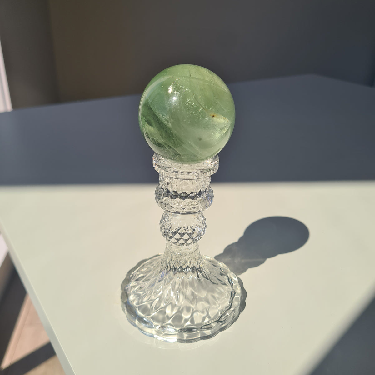 Green fluorite sphere with pale natural bands on a clear decorative stand in sunlight.